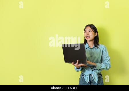 Smiling Young Asian Woman Holding Laptop In Front Of Yellow Background, Business Concept Stock Photo