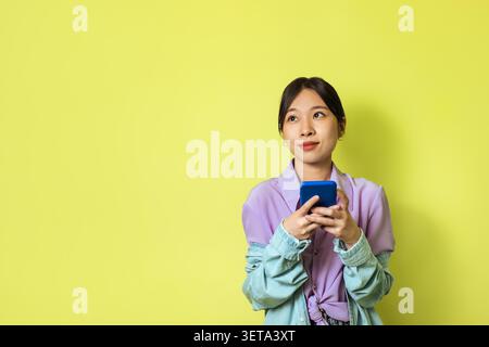 Young Asian Woman Using Smartphone And Smiling, Vibrant Yellow Background Stock Photo