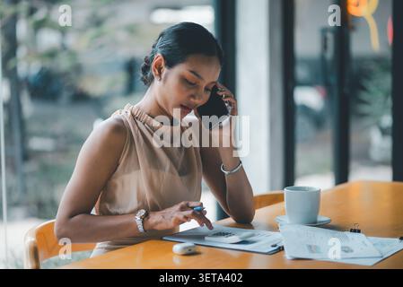 Asian businesswoman talking on smartphone while using calculator and reviewing finance documents in cafe, showing accounting, budgeting, remote work, Stock Photo
