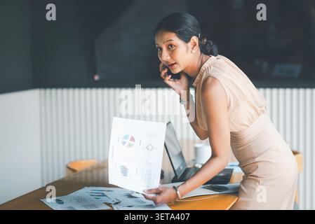 Asian businesswoman talking on smartphone while holding financial report in cafe office, discussing budget results, accounting review, client consulti Stock Photo