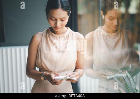 Asian businesswoman standing by glass window using smartphone with reflection, representing mobile finance, digital communication, accounting reminder Stock Photo