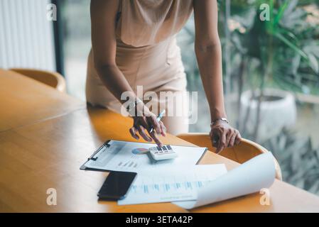 Closeup of businesswoman using calculator on financial report with smartphone and charts at wooden cafe table, showing accounting, budget analysis, pl Stock Photo