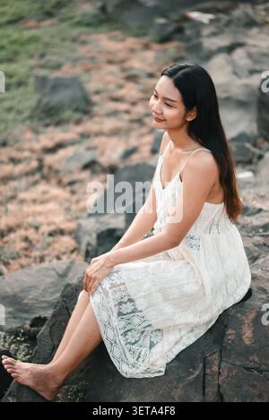 Young Asian woman in a white lace dress sitting on dark rocks in nature, smiling softly with relaxed posture, showing outdoor portrait, peaceful lifes Stock Photo