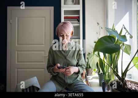 Calm mature woman listening to music in wired headphones using streaming service app on smartphone Stock Photo