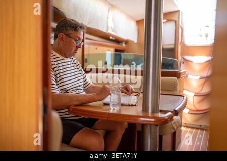 Man working remotely on laptop inside sailboat cabin Stock Photo