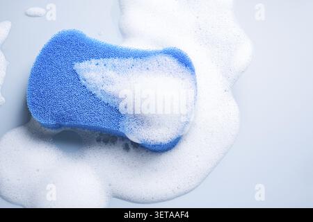 Blue sponge surrounded by white soap foam on a light blue background Stock Photo