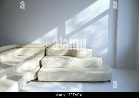 An elegant leather sofa of a pleasant beige color against a white wall. A shadow from the sun falls from the window on a sunny day. The sofa is modern Stock Photo