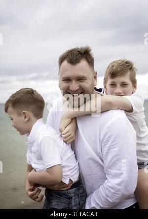 Close up of dad carrying two sons on shoulders on beach. Father and Sons Walking Together on Beach Father and Sons playing and having fun on the beach Stock Photo