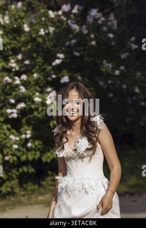 Stunning Bride Gracefully Poses in Spring Park Among Blossoming Trees. Exuding Elegance, Romance and Timeless Charm. long white wedding dress in Europ Stock Photo