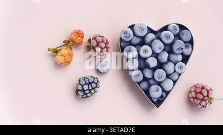 Freshly frozen blueberries in the shape of a metal heart, raspberries, blackberries and cloudberries on a pink background. Summer berry picking. Space Stock Photo
