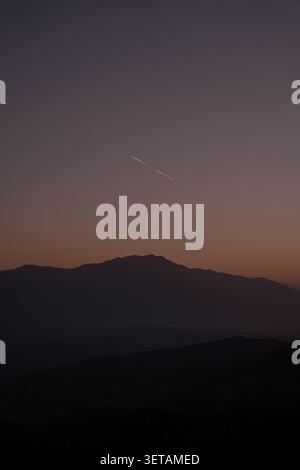 Quiet twilight sky above dark mountain silhouettes with a faint airplane trail, creating a calm scenic landscape with soft purple and orange evening l Stock Photo