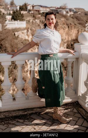 Woman balcony valley posing gracefully in a white blouse and green skirt against a scenic background on a sunny day Stock Photo