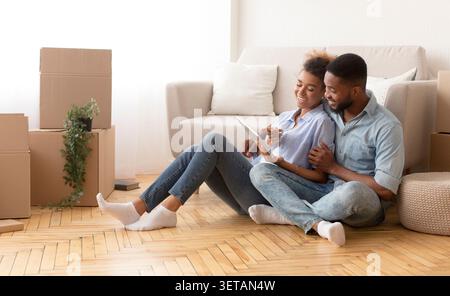 Spouses Using Tablet Sitting On Floor Among Unpacked Boxes Indoor Stock Photo