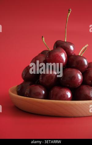 Close-up of dark red cherries spilling from a wooden bowl on a red background, with glossy skin, fresh stems and simple studio lighting. Stock Photo
