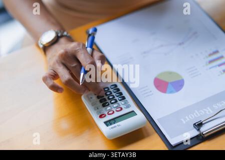 Close-up of a hand pressing a calculator beside a business report with colorful charts, illustrating financial analysis, accounting, budget management Stock Photo