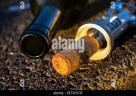 Close-up of an open empty wine bottle lying down with a natural cork and a metal corkscrew on a textured asphalt background during warm golden hour li Stock Photo