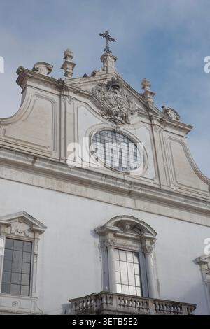 White Baroque Church Facade with Ornate Carved Decoration, Oval Rose Window and Iron Cross Against Blue Sky in Lisbon Stock Photo