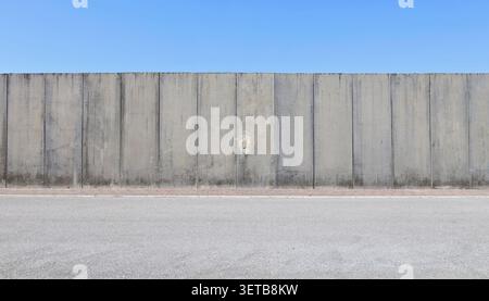 High prefab concrete boundary wall with blue sky above. Red tled sidewalk and street in front. Background for copy space. Stock Photo