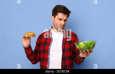 Man Holds Burger in One Hand and Salad in the Other While Showing Uncertainty About His Choice Stock Photo
