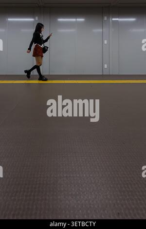 A young Japanese woman walks through Shinjuku Station while using her smartphone. Shinjuku, Tokyo, Japan. Stock Photo