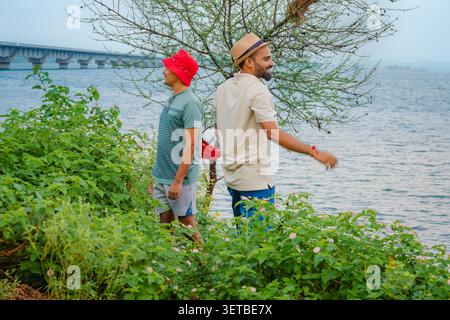 Two men stand near lush green bushes by a large body of water, with a bridge in the distance, Banswara, Udaipur, Rajasthan. Stock Photo