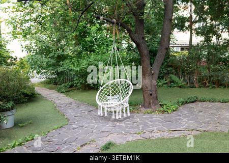 Comfortable hanging wicker white chair in summer garden. Cozy hygge place for weekend relax in garden. Hammock chair in boho style hanging on tree. C Stock Photo