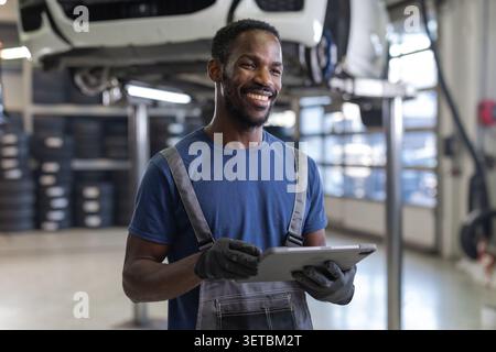 African American mechanic using a tablet to diagnose breakdowns, check maintenance, and order parts, next to lifted cars, portrait. Stock Photo