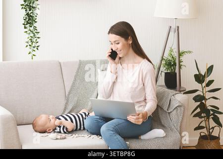 Mother Talking on Phone While Using Laptop With Baby on Couch in Living Room Stock Photo
