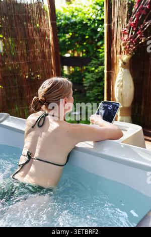 Outdoor spa lifestyle concept with woman using digital hot tub controls while enjoying hydrotherapy in a private garden setting. Stock Photo