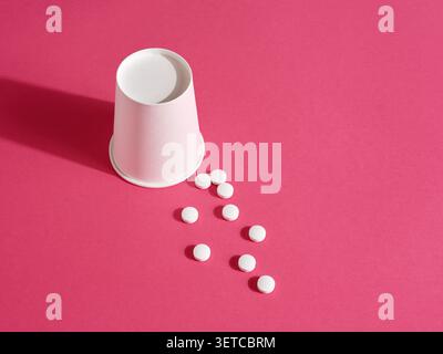 White pills spilling from a disposable paper cup on a vibrant pink background, creating a simple yet striking image related to medicine, healthcare, a Stock Photo