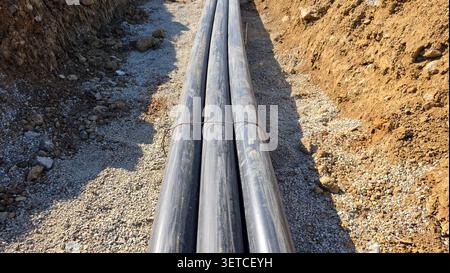Three black industrial protective pipelines are secured together by a metal wire tie, resting on a bed of crushed gravel within an open earthen Stock Photo
