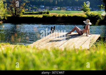 Woman in swimsuit, tourist sitting on a wooden pier on Krka river on a sunny hot day in Dolenjska region in Slovenia, summer holidays Stock Photo