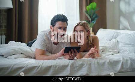 Joyful Caucasian couple using mobile phone laughing in bed family watching funny humorous video enjoy weekend lying in bedroom man with woman watch Stock Photo