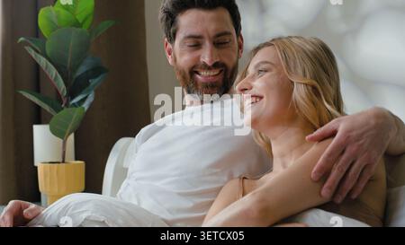 Happy family relaxed Caucasian couple cuddle talking communicating relaxing lying in bed cozy morning at home woman cuddling with lovely man talk Stock Photo