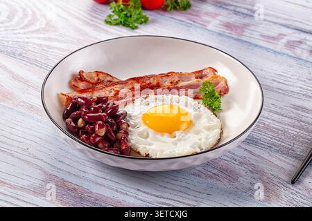 Appetizing breakfast plate features sunny side up egg, crispy bacon strips, red kidney beans. White bowl on light rustic wooden table Stock Photo