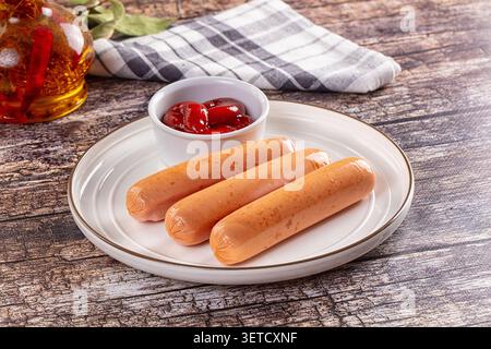 Three golden brown sausages served on white ceramic plate with small bowl of red ketchup. Wooden rustic table background with checkered napkin chili o Stock Photo