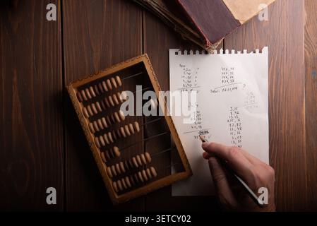 Top view of an accountant hand calculating budget expenses on paper with a pen, next to old wooden abacus and books on table. Stock Photo