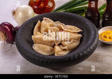 Homemade vareniki dumplings served in rustic black stone bowl with red onion, white onion, green onions, mustard sauce, spices on light background. Tr Stock Photo