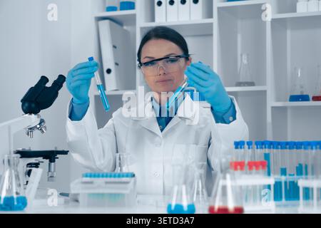 In a wellequipped lab, a focused female scientist in protective gear examines two test tubes with different substances, surrounded by glassware and advanced equipment for her research Stock Photo