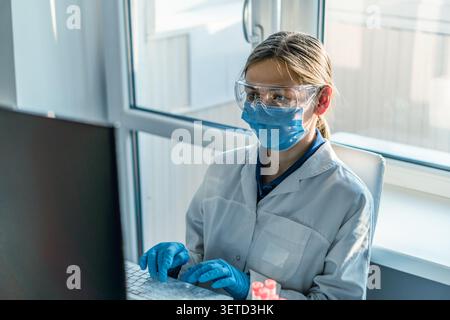 Young woman scientist wearing lab coat, face mask, gloves, and safety goggles sits at desk with computer in bright laboratory. She is conducting scientific research and analyzing experimental results Stock Photo