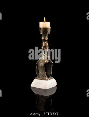 Artistic bronze candle holder sculpture by artist Lukas with a lit candle on a white marble base, photographed on a black background. Stock Photo
