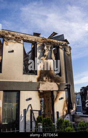 A medium-shot view of a modern multi-story residential building or townhouse heavily damaged by a catastrophic fire or an explosion. The upper floor i Stock Photo