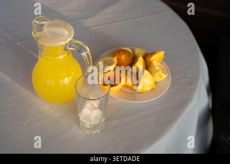 Clear glass pitcher filled with bright yellow lemonade placed on round white tablecloth, beside tall empty glass and plate piled with halved and sliced lemon Stock Photo
