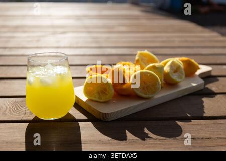 Glass of pale yellow lemon juice with ice placed on sunlit wooden table next to wooden cutting board holding halved and squeezed lemons, warm natural light Stock Photo
