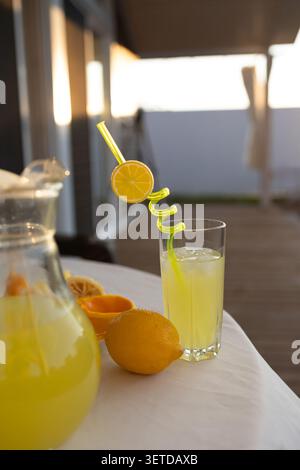 Tall glass of pale yellow lemonade with ice and decorative curvy straw placed on white tablecloth beside whole and halved lemons and pitcher, set on wooden Stock Photo