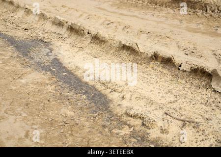 Deep tire tracks are imprinted on a dry mud road surface. Textured car wheel tread marks cover the bumpy clay and sandy ground path of a rural country Stock Photo