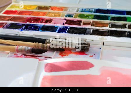 Artist watercolor paints palette and wet brushes lie near a sketchbook. Colorful aquarelle pigments box contains diverse wet paint pans with textured Stock Photo