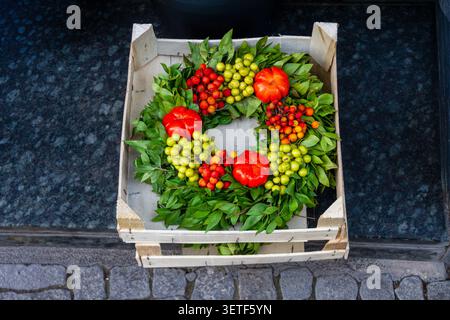 A decorative green leaf wreath is arranged inside a rustic wooden crate on a dark stone step. The arrangement is enriched with three small bumpy red p Stock Photo