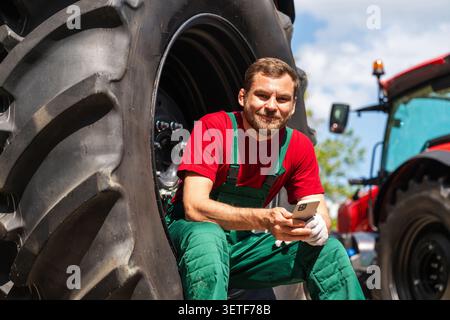 Adult male farmer with phone sitting in big wheel of tractor at farm. Stock Photo