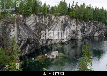Towering gray rock walls rise over clear green water beside dense pine forest, with tiny visitors on platforms and raft near shoreline Stock Photo